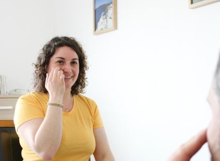Femme souriante en haut jaune, gesticulant dans une pièce lumineuse.
